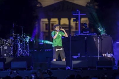 A concert scene features a performer in a green shirt and dark shorts energetically singing into a microphone. The stage is illuminated with colorful lights, and musical equipment including drums and a DJ setup are visible. In the background, a large building can be seen.