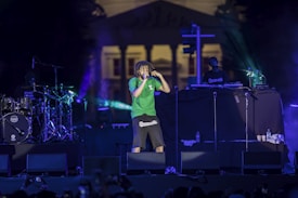 A concert scene features a performer in a green shirt and dark shorts energetically singing into a microphone. The stage is illuminated with colorful lights, and musical equipment including drums and a DJ setup are visible. In the background, a large building can be seen.