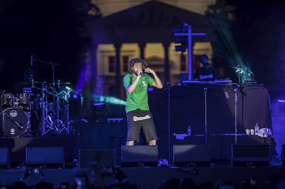 A concert scene features a performer in a green shirt and dark shorts energetically singing into a microphone. The stage is illuminated with colorful lights, and musical equipment including drums and a DJ setup are visible. In the background, a large building can be seen.