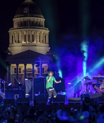 A live outdoor concert is taking place at night with a grand, dome-shaped architectural structure illuminated in the background. On stage, a person wearing a green shirt and black shorts is energetically performing. Colorful stage lights in blue, purple, and green add vibrance to the scene, and other band members or crew can be seen to the sides. The audience is visible in the foreground.