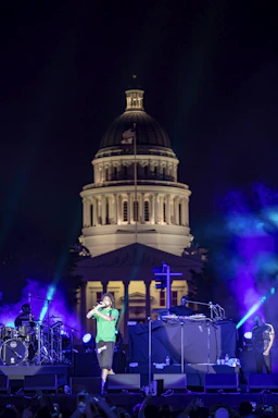 A live music performance takes place in front of an illuminated, historic-looking building with a large dome. The stage is lit with vibrant purple and blue lights, and a performer in a green shirt and black shorts is singing or speaking into a microphone. There are musicians and equipment visible on the stage, including a drum set and turntables.