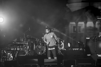 A performer stands on stage holding a microphone, surrounded by musical instruments like a drum set and a keyboard. The lighting is dramatic with bright stage lights creating a contrast with the dark background. The scene appears to be an energetic live performance.