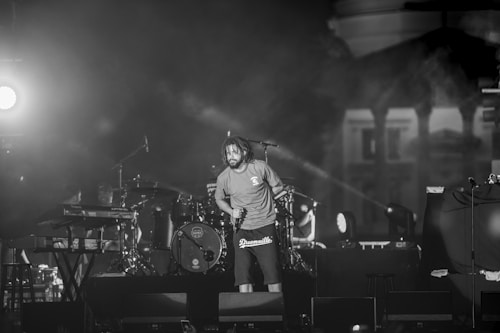 A performer stands on stage holding a microphone, surrounded by musical instruments like a drum set and a keyboard. The lighting is dramatic with bright stage lights creating a contrast with the dark background. The scene appears to be an energetic live performance.