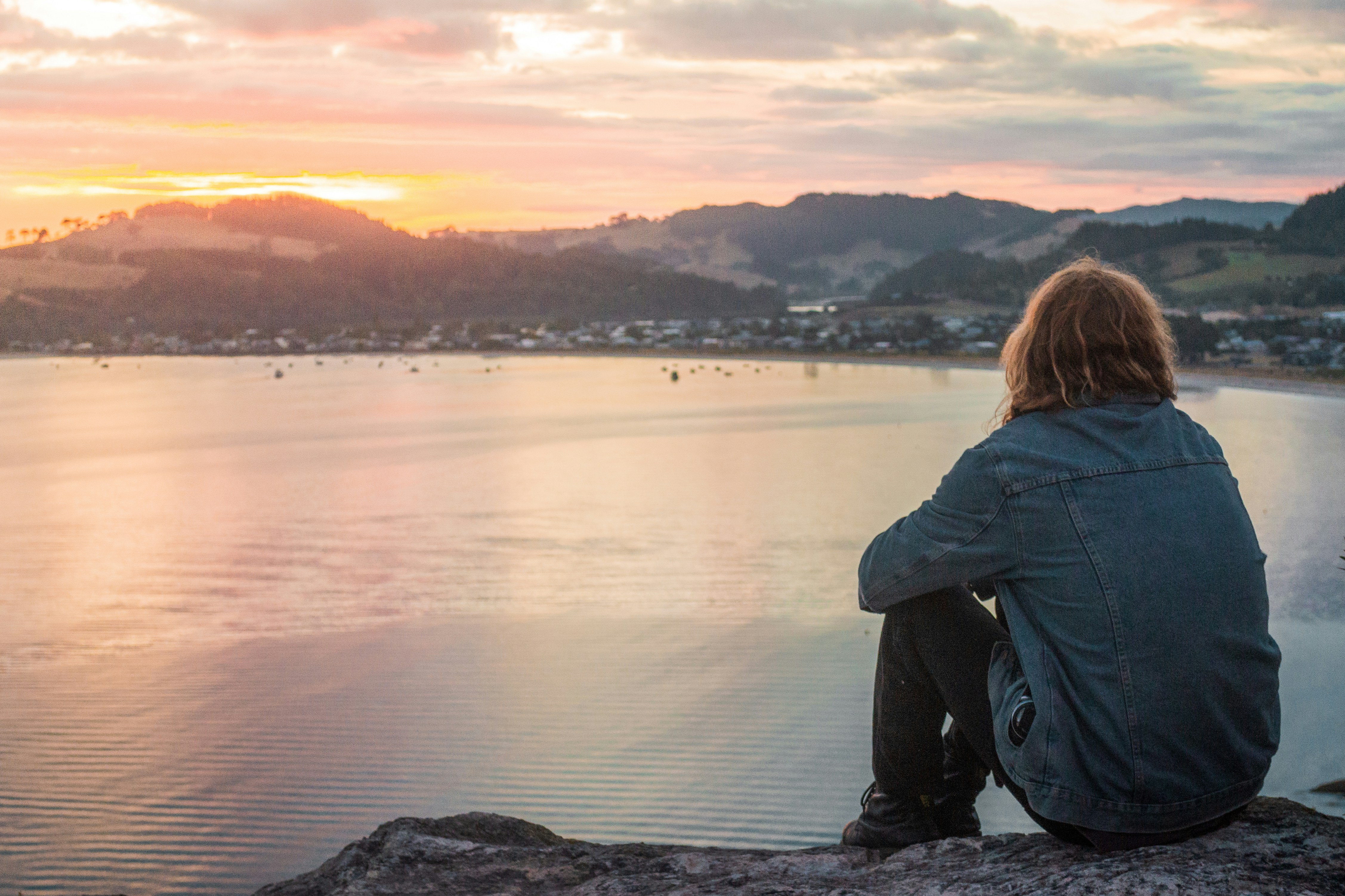 woman in blue denim jacket sitting on rock near body of water during sunset
