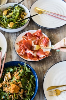 The image features a spread of dishes on a wooden table. There are plates of prosciutto and arugula salad, with some containing grains and vegetables. Gold and black cutlery accompanies the plates, and a hand is using a fork to pick up the prosciutto.