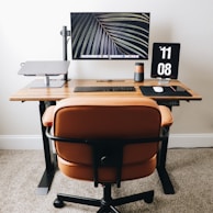 A modern workspace setup featuring a wooden desk with a large monitor displaying a leaf pattern, a laptop elevated on a stand, a digital clock showing the time as 11:08, a wireless keyboard and mouse, and a smartphone on a black mouse pad. An ergonomic brown leather chair is positioned in front of the desk, and the floor is carpeted.