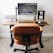 A modern workspace setup featuring a wooden desk with a large monitor displaying a leaf pattern, a laptop elevated on a stand, a digital clock showing the time as 11:08, a wireless keyboard and mouse, and a smartphone on a black mouse pad. An ergonomic brown leather chair is positioned in front of the desk, and the floor is carpeted.
