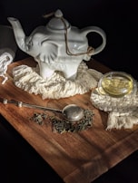 A ceramic teapot shaped like an elephant sits on a wooden surface, surrounded by a woven mat and a glass cup filled with tea. A metal tea infuser and loose tea leaves are scattered nearby, creating an inviting tea setup illuminated by soft light.
