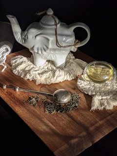 A ceramic teapot shaped like an elephant sits on a wooden surface, surrounded by a woven mat and a glass cup filled with tea. A metal tea infuser and loose tea leaves are scattered nearby, creating an inviting tea setup illuminated by soft light.