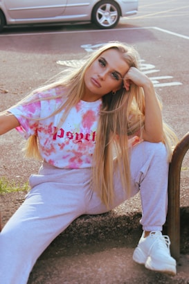 A person with long blonde hair is sitting on a pavement, posing with a relaxed expression. They are wearing a tie-dye t-shirt with the word 'psych' printed on it, gray sweatpants, and white sneakers. One arm is resting on their knee while the other supports their head. A parked car is visible in the background.