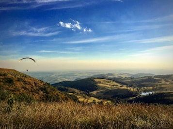 A picturesque landscape featuring a paraglider soaring above rolling hills covered with a mix of grassland and patches of forest. The sky is clear and blue with a few scattered clouds, casting gentle shadows on the terrain. A small water body is visible in the distance, reflecting the sunlight.