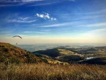 A picturesque landscape featuring a paraglider soaring above rolling hills covered with a mix of grassland and patches of forest. The sky is clear and blue with a few scattered clouds, casting gentle shadows on the terrain. A small water body is visible in the distance, reflecting the sunlight.