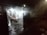 A night scene depicting a gathering in a dimly lit outdoor area under a roofed patio. Several people are standing and sitting around a table with food, and there's visible smoke possibly from a barbecue or cooking activity. The area is illuminated by overhead lights, and the surrounding is dark and slightly wet, suggesting a recent rain.