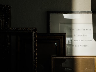 Several picture frames are stacked against a wall, creating a layered arrangement. A frame on the right contains a motivational quote that reads 'Whatever the mind of man can conceive and believe, it can achieve.' A shadow partially covers the frames, adding depth and contrast.