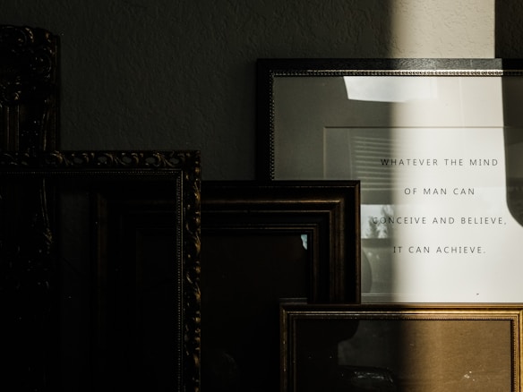 Several picture frames are stacked against a wall, creating a layered arrangement. A frame on the right contains a motivational quote that reads 'Whatever the mind of man can conceive and believe, it can achieve.' A shadow partially covers the frames, adding depth and contrast.