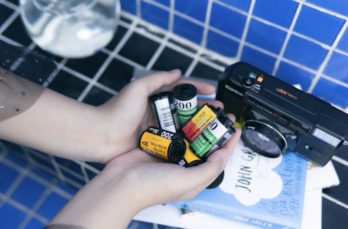 A pair of hands gently holding multiple rolls of photographic film, with a vintage Konica camera placed beside them on a table covered with a blue-tiled pattern. In the background, a couple of books are visible with text on the cover.