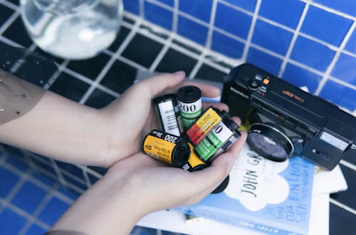 A pair of hands gently holding multiple rolls of photographic film, with a vintage Konica camera placed beside them on a table covered with a blue-tiled pattern. In the background, a couple of books are visible with text on the cover.