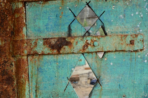 A weathered and rusted metal surface features a turquoise paint with large patches of rust. The surface includes a geometric pattern with diamond-shaped cutouts, revealing a slightly blurred view of an exterior background.
