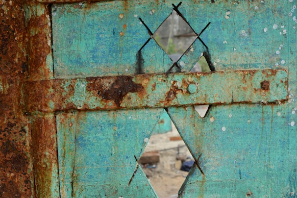 A weathered and rusted metal surface features a turquoise paint with large patches of rust. The surface includes a geometric pattern with diamond-shaped cutouts, revealing a slightly blurred view of an exterior background.