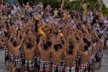 A cultural performance is taking place with a large group of people forming a circle. A performer in the center is dressed in traditional colorful attire and adorned with an ornate headdress. The crowd surrounding them is engaged, raising their arms and appearing to participate actively. The audience watches intently in the background.