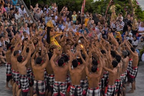 A cultural performance is taking place with a large group of people forming a circle. A performer in the center is dressed in traditional colorful attire and adorned with an ornate headdress. The crowd surrounding them is engaged, raising their arms and appearing to participate actively. The audience watches intently in the background.