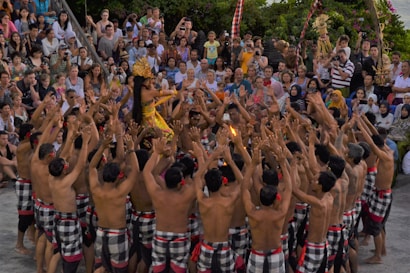 A cultural performance is taking place with a large group of people forming a circle. A performer in the center is dressed in traditional colorful attire and adorned with an ornate headdress. The crowd surrounding them is engaged, raising their arms and appearing to participate actively. The audience watches intently in the background.