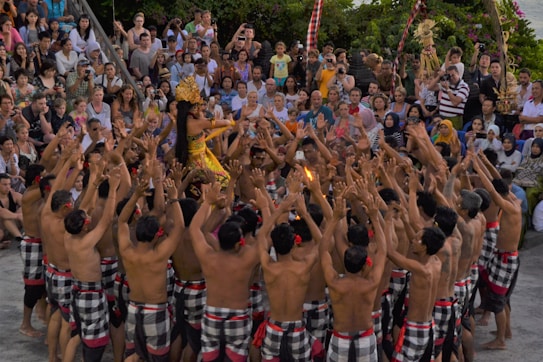 A cultural performance is taking place with a large group of people forming a circle. A performer in the center is dressed in traditional colorful attire and adorned with an ornate headdress. The crowd surrounding them is engaged, raising their arms and appearing to participate actively. The audience watches intently in the background.