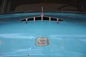 A close-up view of the front part of a vintage car in a bright blue color, showcasing a metallic vent and the manufacturer's emblem labeled 'Austin'. The surface appears glossy with visible reflections.