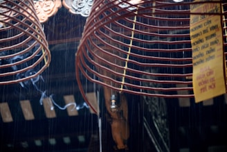 A close-up of two large red spiral incense coils hanging from above with a thin trail of smoke rising from them. There are intricate decorative elements at the top and a yellow sign with text in a foreign language attached to one of the coils. The background appears to be part of a dimly lit wooden or metal structure, possibly a temple or shrine.