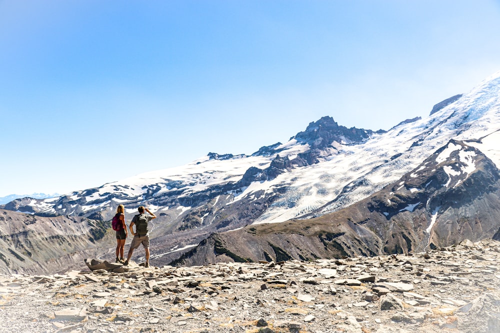 2 person standing on rocky ground near snow covered mountain during daytime
