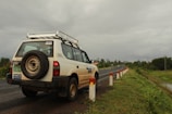 A white SUV is parked on the side of a road, with a visible spare tire mounted on the back. The road is flanked by a series of white and red posts, and it stretches into the distance. The surroundings are lush with greenery, and the sky is overcast with a heavy, gray cloud cover.