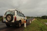 A white SUV is parked on the side of a road, with a visible spare tire mounted on the back. The road is flanked by a series of white and red posts, and it stretches into the distance. The surroundings are lush with greenery, and the sky is overcast with a heavy, gray cloud cover.