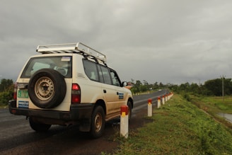 A white SUV is parked on the side of a road, with a visible spare tire mounted on the back. The road is flanked by a series of white and red posts, and it stretches into the distance. The surroundings are lush with greenery, and the sky is overcast with a heavy, gray cloud cover.