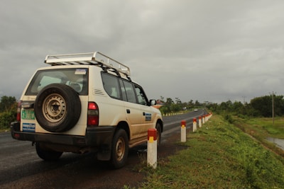 A white SUV is parked on the side of a road, with a visible spare tire mounted on the back. The road is flanked by a series of white and red posts, and it stretches into the distance. The surroundings are lush with greenery, and the sky is overcast with a heavy, gray cloud cover.