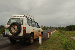 A white SUV is parked on the side of a road, with a visible spare tire mounted on the back. The road is flanked by a series of white and red posts, and it stretches into the distance. The surroundings are lush with greenery, and the sky is overcast with a heavy, gray cloud cover.