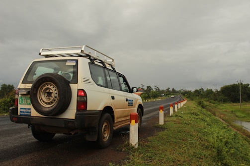 A white SUV is parked on the side of a road, with a visible spare tire mounted on the back. The road is flanked by a series of white and red posts, and it stretches into the distance. The surroundings are lush with greenery, and the sky is overcast with a heavy, gray cloud cover.
