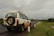 A white SUV is parked on the side of a road, with a visible spare tire mounted on the back. The road is flanked by a series of white and red posts, and it stretches into the distance. The surroundings are lush with greenery, and the sky is overcast with a heavy, gray cloud cover.