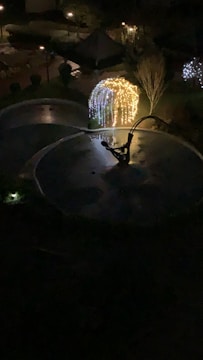 A nighttime scene featuring a garden area illuminated by decorative string lights arranged in a circular arch. The lights form a glowing structure against the dark surroundings. A water feature or fountain with reflective surfaces is also visible, adding to the serene ambiance.