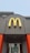 A McDonald's sign with a yellow 'M' logo and a red maple leaf located above the entrance of a building. The building features grey walls with bright orange accents. Below the sign, glass doors display a promotion for summer drink days.
