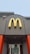 A McDonald's sign with a yellow 'M' logo and a red maple leaf located above the entrance of a building. The building features grey walls with bright orange accents. Below the sign, glass doors display a promotion for summer drink days.