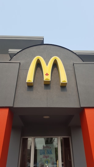 A McDonald's sign with a yellow 'M' logo and a red maple leaf located above the entrance of a building. The building features grey walls with bright orange accents. Below the sign, glass doors display a promotion for summer drink days.