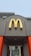 A McDonald's sign with a yellow 'M' logo and a red maple leaf located above the entrance of a building. The building features grey walls with bright orange accents. Below the sign, glass doors display a promotion for summer drink days.