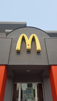 A McDonald's sign with a yellow 'M' logo and a red maple leaf located above the entrance of a building. The building features grey walls with bright orange accents. Below the sign, glass doors display a promotion for summer drink days.