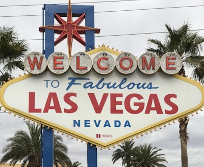 A large, iconic sign reads 'Welcome to Fabulous Las Vegas Nevada' with vintage-style lettering. The sign is topped with a red star and flanked by palm trees against a cloudy sky.