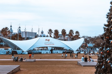 A large, white, tent-like structure serves as the focal point in a park-like setting with several people gathered around. The structure is adorned with signage for the Tampa Bay Buccaneers Foundation and Winter Village. In the background, palm trees and ornate historic buildings are visible. On one side, a decorated Christmas tree stands.