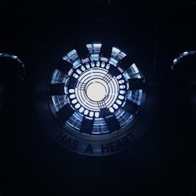 A circular, industrial-looking structure with concentric rings emits a bright white light from the center. Surrounding the center, there are several black radial elements, resembling a mechanical or technological device. The words 'HAS A HEART' are partially visible along the outer edge.