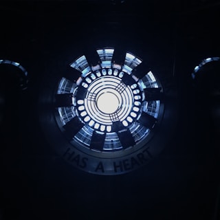 A circular, industrial-looking structure with concentric rings emits a bright white light from the center. Surrounding the center, there are several black radial elements, resembling a mechanical or technological device. The words 'HAS A HEART' are partially visible along the outer edge.