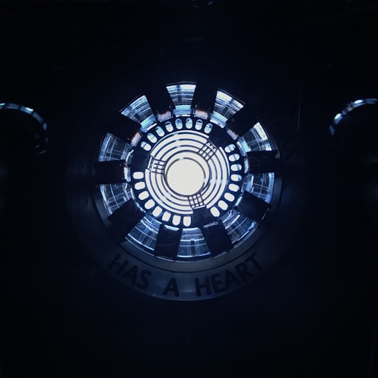 A circular, industrial-looking structure with concentric rings emits a bright white light from the center. Surrounding the center, there are several black radial elements, resembling a mechanical or technological device. The words 'HAS A HEART' are partially visible along the outer edge.
