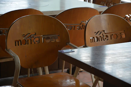 A set of wooden chairs with cut-out lettering on the backrests can be seen. The chairs are arranged around wooden tables, suggesting a cafe or restaurant setting. The lighting is soft and natural, creating subtle reflections on the surfaces.