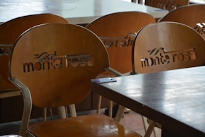 A set of wooden chairs with cut-out lettering on the backrests can be seen. The chairs are arranged around wooden tables, suggesting a cafe or restaurant setting. The lighting is soft and natural, creating subtle reflections on the surfaces.